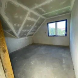 Poddasze po szpachlowaniu, gotowe do malowania. Widoczne gładzie na skosach i ścianach, okno zabezpieczone folią malarską, surowa podłoga. Drewniana listwa w lewym dolnym rogu.