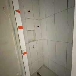 Fachowe Usługi Remontowe Dawid Czosnek - Wnętrze kabiny prysznicowej po wyłożeniu białymi płytkami. Widoczne fragmenty niedokończonej ściany z instalacją. Na płytkach widoczne czerwone i niebieskie znaczniki.