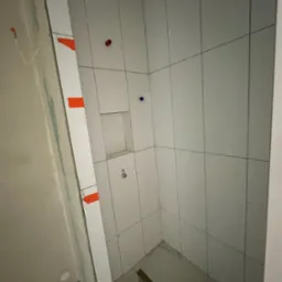 Wnętrze kabiny prysznicowej po wyłożeniu białymi płytkami. Widoczne fragmenty niedokończonej ściany z instalacją. Na płytkach widoczne czerwone i niebieskie znaczniki.