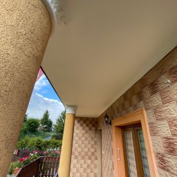 Fachowe Usługi Remontowe Dawid Czosnek - Wykończony balkon z kolumnami i dekoracyjnymi sztukateriami, widok z dołu na sufit i fragment otoczenia. Zewnętrzna elewacja z cegieł.