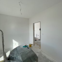 Fachowe Usługi Remontowe Dawid Czosnek - Wnętrze po malowaniu ścian na biało, z zabezpieczonymi podłogami folią. Widoczny kabel zwisający z sufitu, drabina i przejście do kolejnego pomieszczenia.