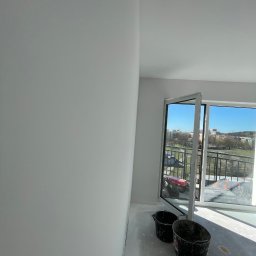 Fachowe Usługi Remontowe Dawid Czosnek - Świeżo pomalowane na biało ściany w pokoju z otwartym oknem balkonowym i dwoma czarnymi wiadrami na podłodze. Widok na miasto w tle.