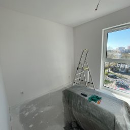 Fachowe Usługi Remontowe Dawid Czosnek - Białe, odświeżone wnętrze pokoju po malowaniu. Widoczna drabina, zabezpieczone meble folią, okno z widokiem na miasto i kabel wystający z sufitu.