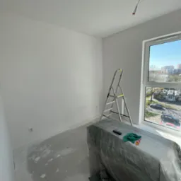 Białe, odświeżone wnętrze pokoju po malowaniu. Widoczna drabina, zabezpieczone meble folią, okno z widokiem na miasto i kabel wystający z sufitu.