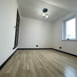 Fachowe Usługi Remontowe Dawid Czosnek - Jasne panele podłogowe w nowym pokoju z białymi ścianami i oknem. Minimalistyczna lampa sufitowa, ciemne listwy przypodłogowe.
