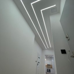 Fachowe Usługi Remontowe Dawid Czosnek - Nowoczesne oświetlenie sufitowe LED w kształcie zygzaka w korytarzu, widok na białą kuchnię w tle. Minimalistyczny design, białe ściany, jasne drewno na podłodze.