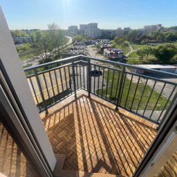 Fachowe Usługi Remontowe Dawid Czosnek - Jasne drewniane płytki na balkonie z metalową balustradą i widokiem na miasto w słoneczny dzień. Charakterystyczne cienie rzucane przez balustradę na podłogę.