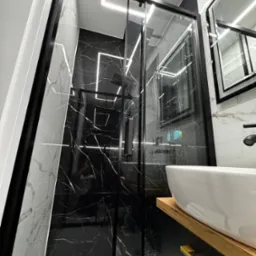 Nowoczesna łazienka z kabiną prysznicową typu walk-in, czarnymi płytkami imitującymi marmur i białym blatem umywalki z drewnianym akcentem. Oświetlenie LED.