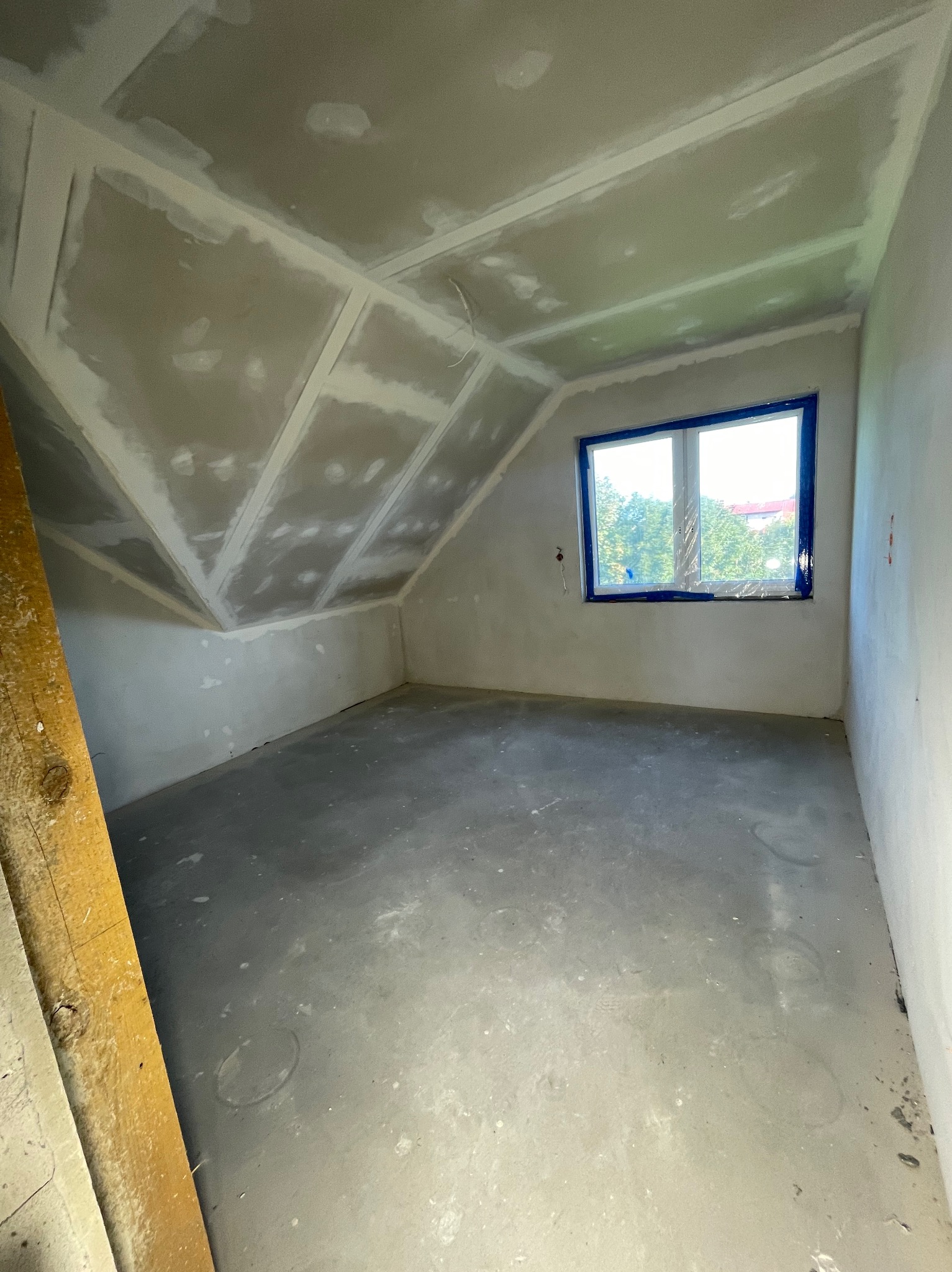 Poddasze po szpachlowaniu, gotowe do malowania. Widoczne gładzie na skosach i ścianach, okno zabezpieczone folią malarską, surowa podłoga. Drewniana listwa w lewym dolnym rogu.