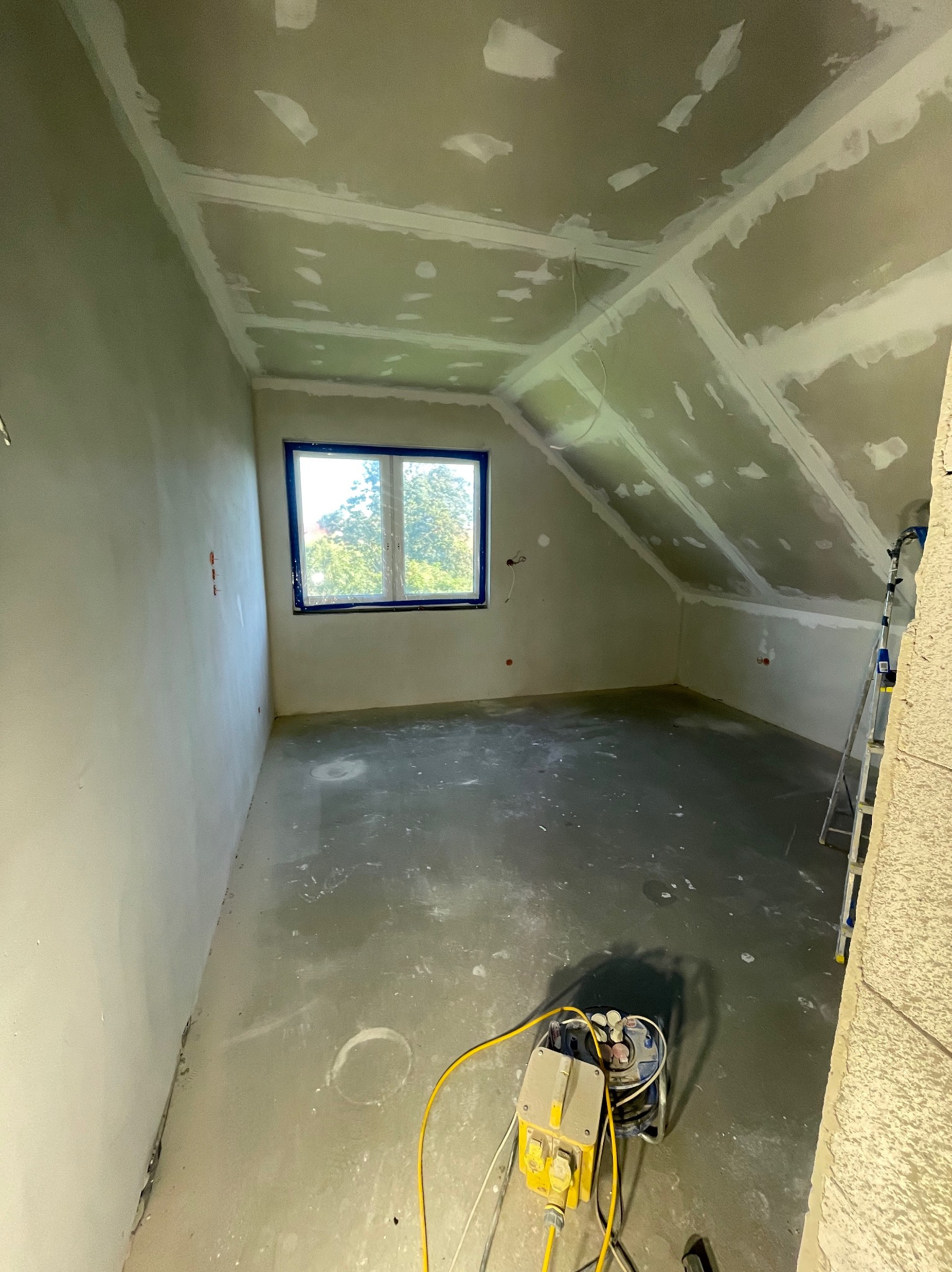 Poddasze po szpachlowaniu w Grzybnie: widok na ścianę, sufit ze śladami po łatach i okno. Na podłodze leży maszyna do szpachlowania, a w rogu stoi drabina.
