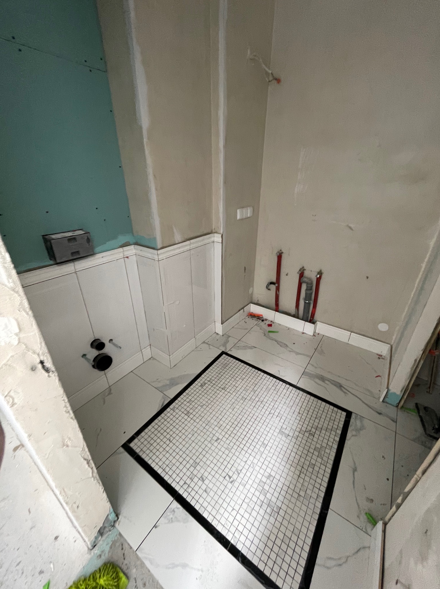 Wnętrze łazienki w trakcie prac: białe płytki na ścianach do połowy, szara ściana powyżej, mozaika na podłodze z czarną obwódką, instalacja wodna.