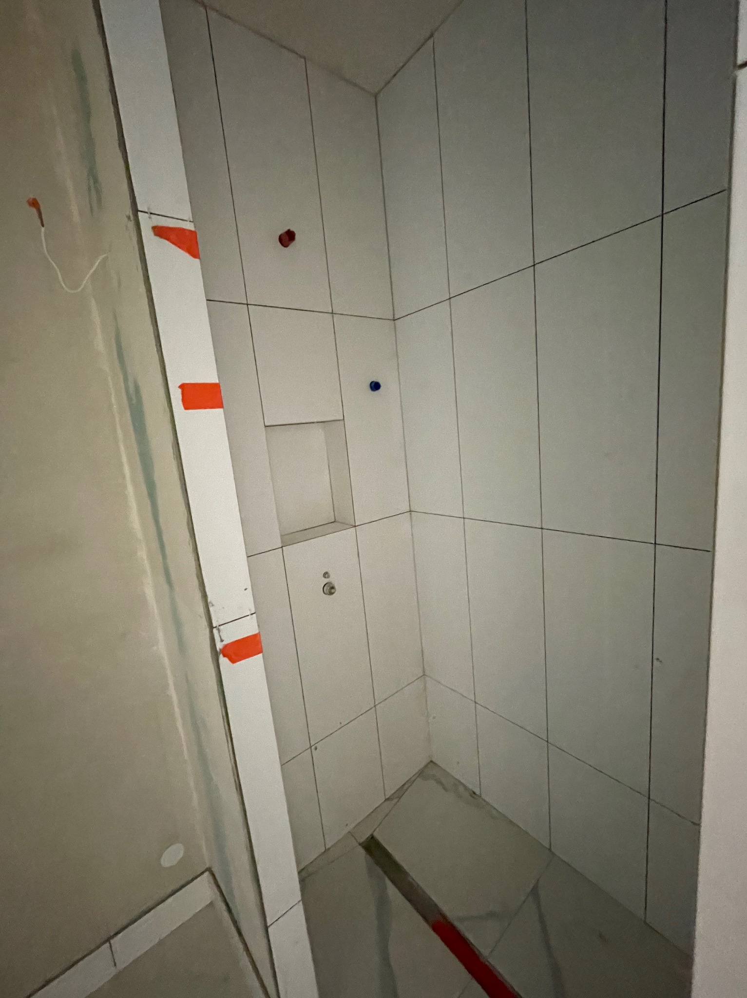 Wnętrze kabiny prysznicowej po wyłożeniu białymi płytkami. Widoczne fragmenty niedokończonej ściany z instalacją. Na płytkach widoczne czerwone i niebieskie znaczniki.