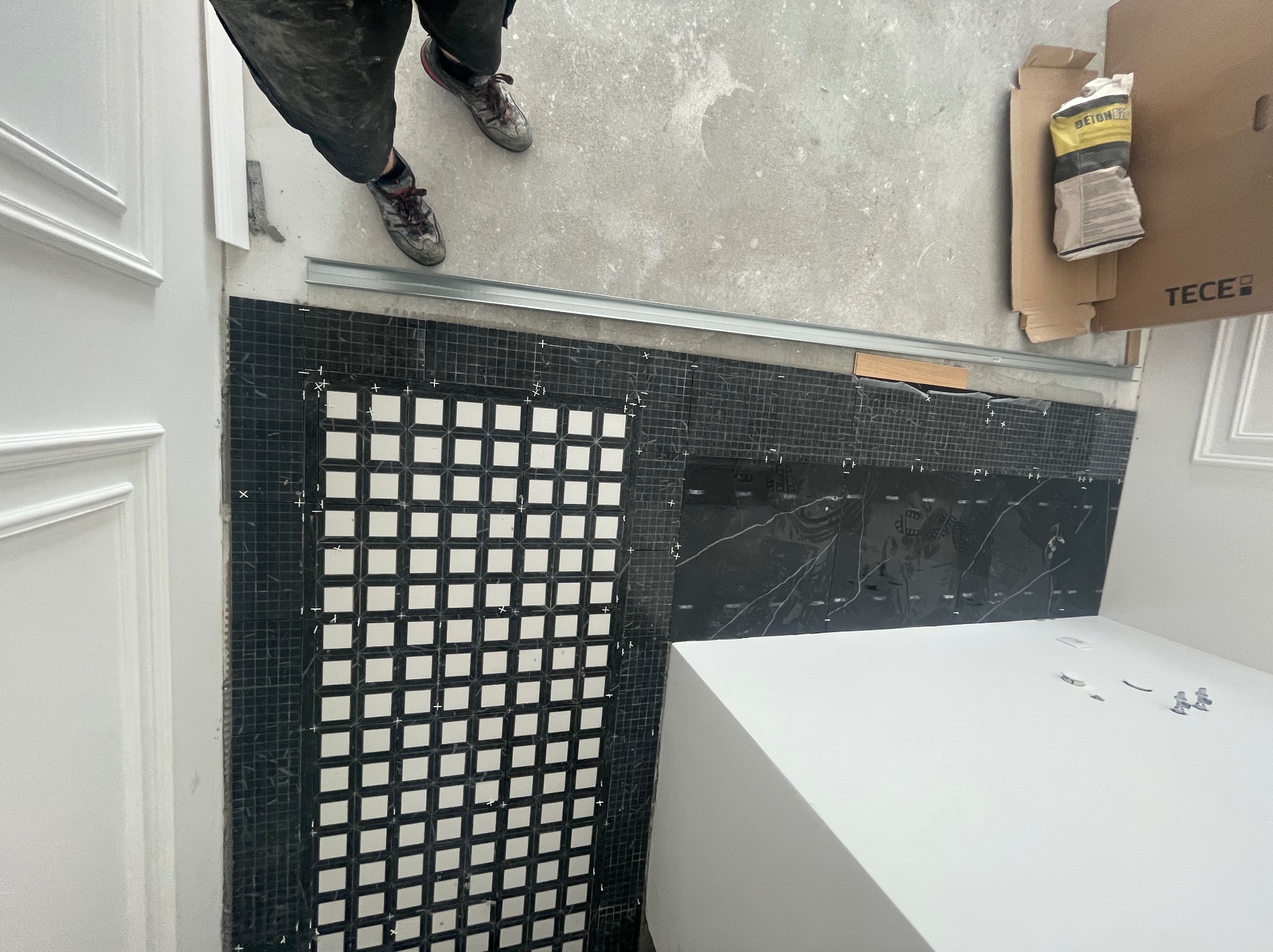 Układanie mozaiki w łazience: czarno-biała szachownica przy białej wannie, widok z góry, widoczne narzędzia i worek z zaprawą. Etap prac wykończeniowych.