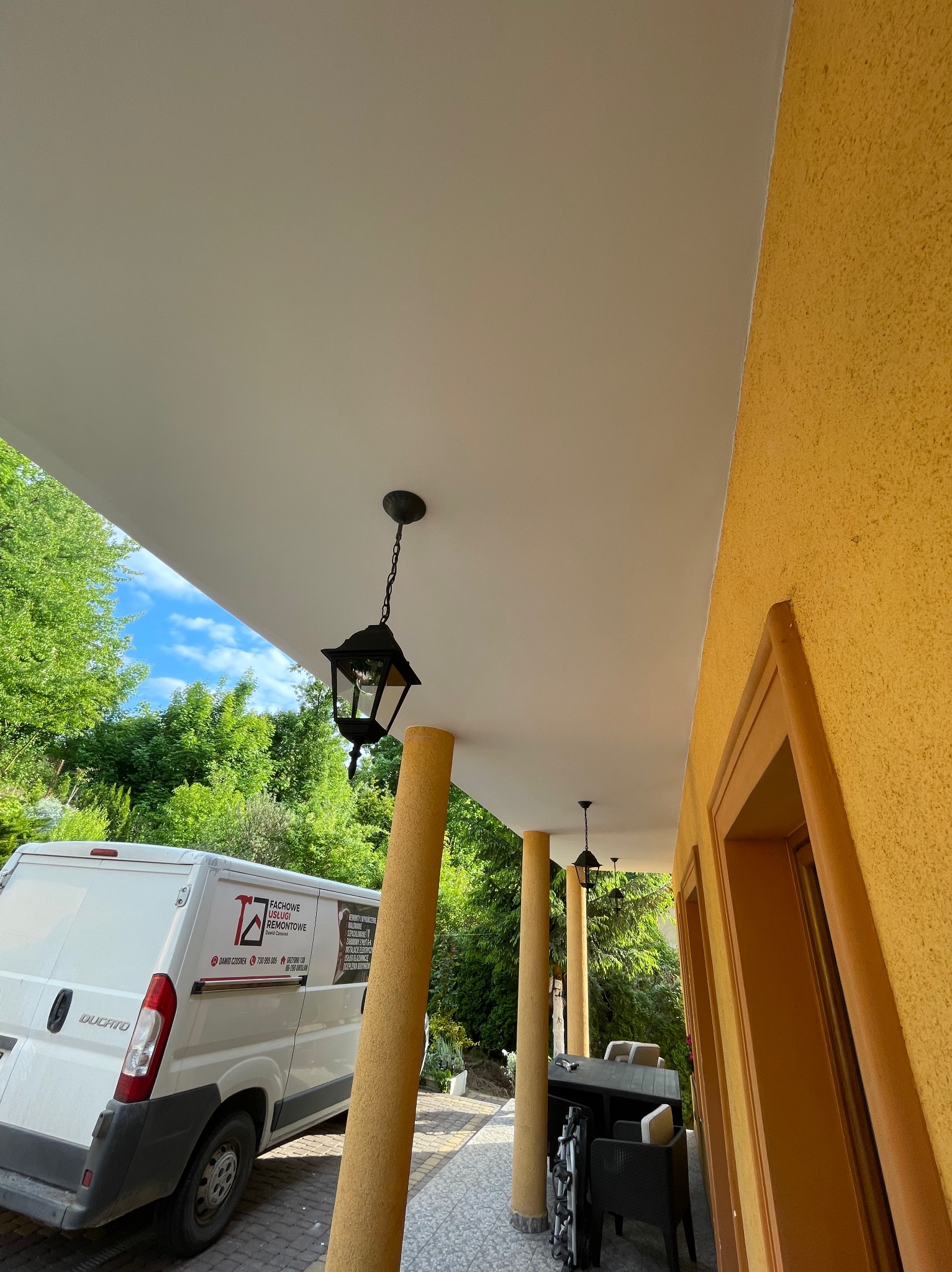 Zadaszony taras z oświetleniem i widoczną elewacją domu w odcieniach żółci. W tle zieleń drzew i zaparkowany biały bus z logo firmy remontowej.