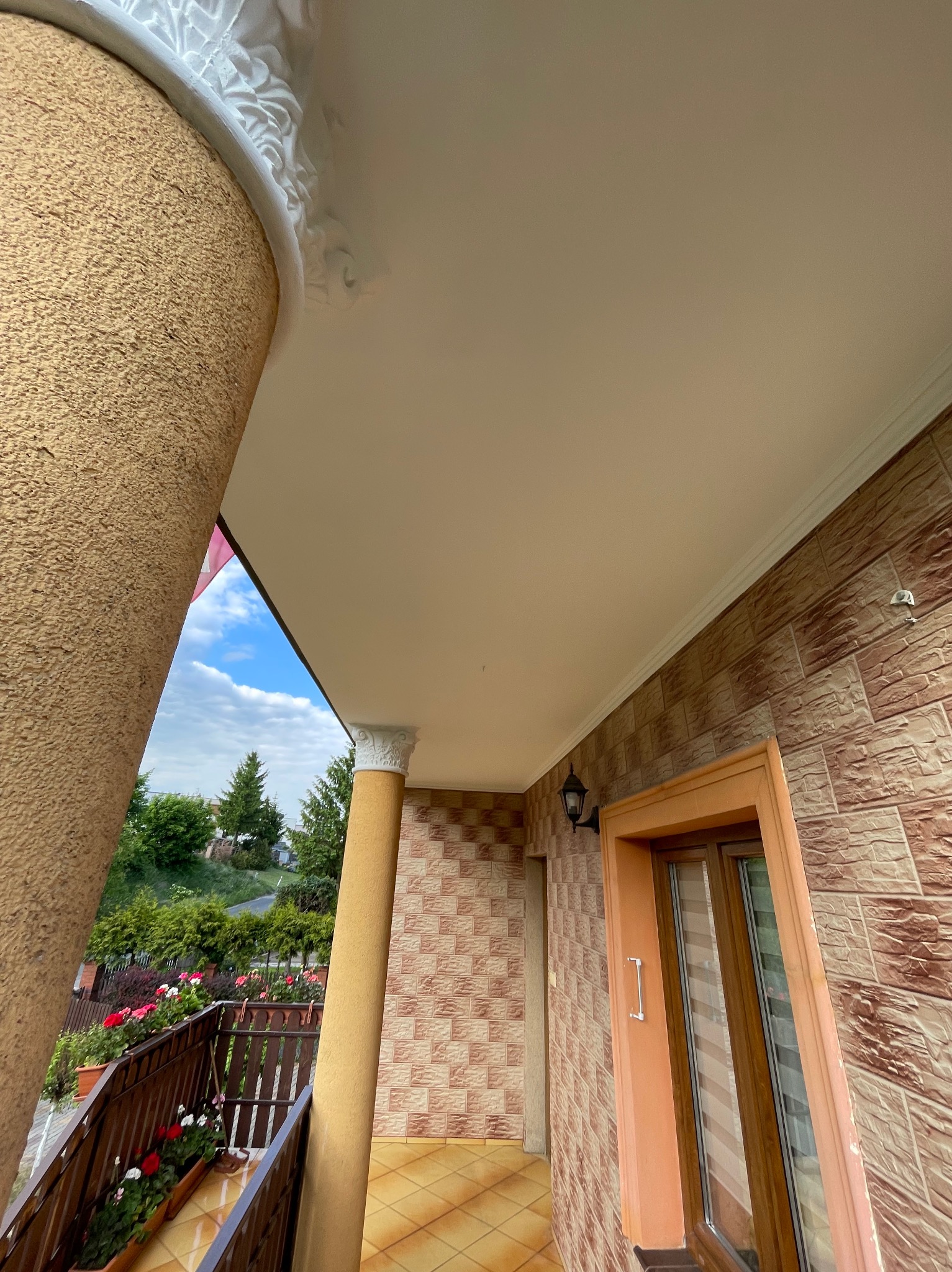 Wykończony balkon z kolumnami i dekoracyjnymi sztukateriami, widok z dołu na sufit i fragment otoczenia. Zewnętrzna elewacja z cegieł.