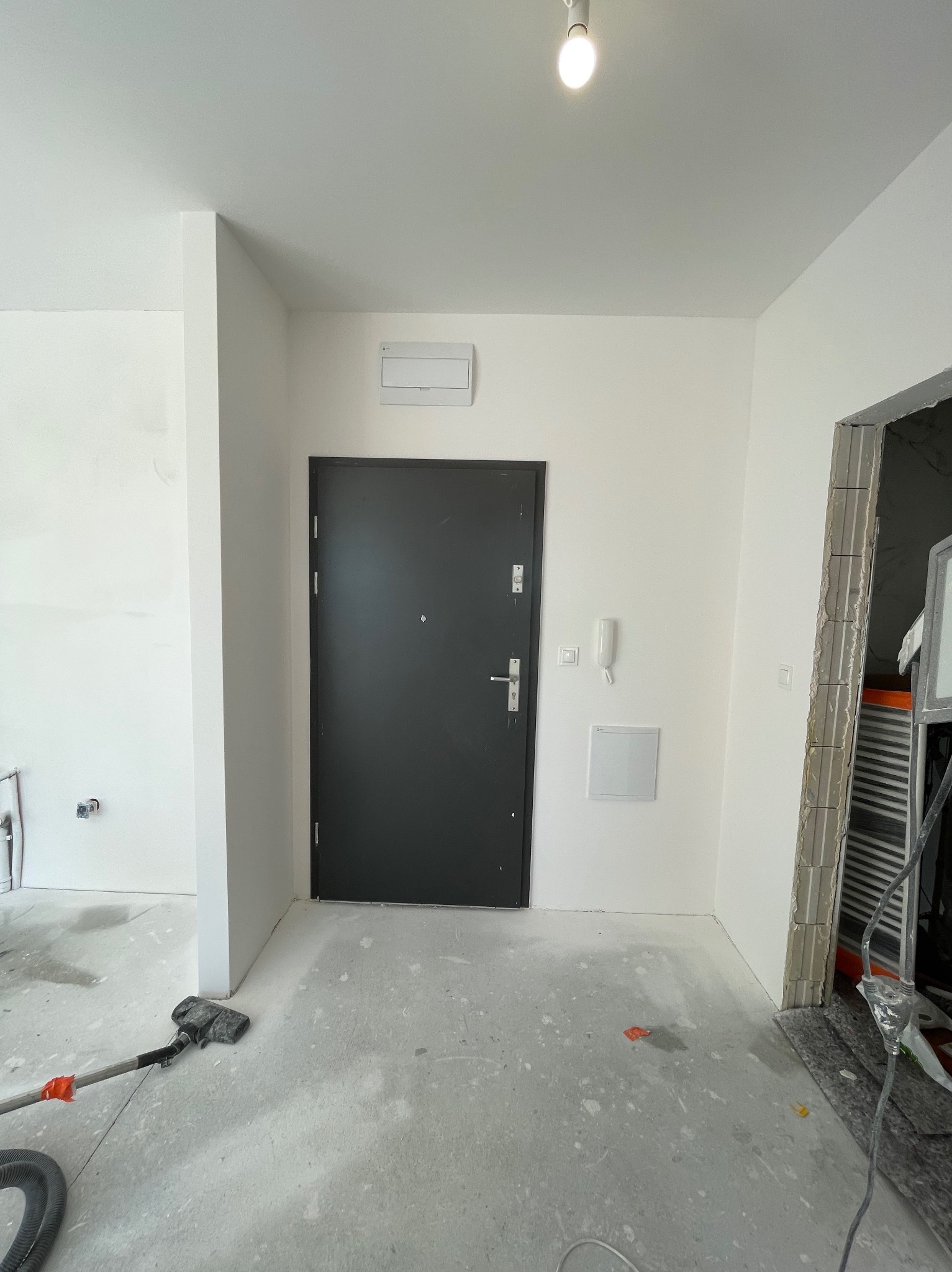 Wnętrze w trakcie prac wykończeniowych: szare drzwi wejściowe, białe ściany po szpachlowaniu, widoczny odkurzacz przemysłowy i fragmenty prac murarskich przy ościeżnicy.