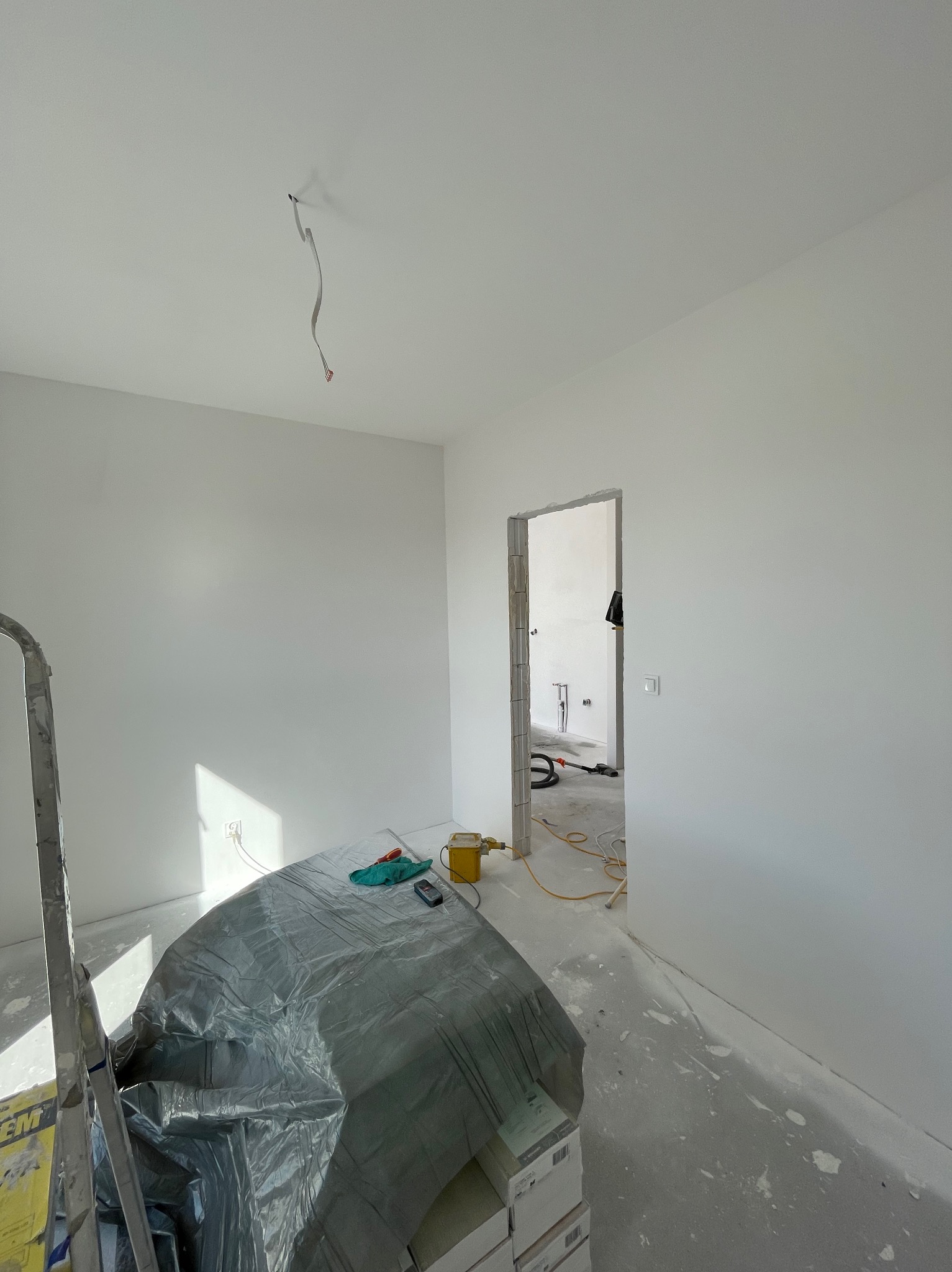 Wnętrze po malowaniu ścian na biało, z zabezpieczonymi podłogami folią. Widoczny kabel zwisający z sufitu, drabina i przejście do kolejnego pomieszczenia.