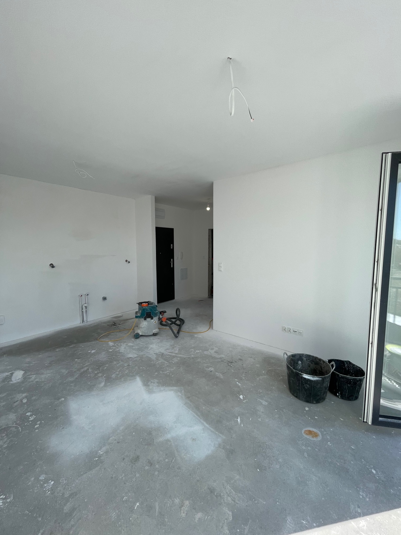 Wnętrze w trakcie prac: białe ściany, surowa podłoga, narzędzia (szlifierka, wiadra). Widoczne instalacje i kable. Minimalistyczny styl, surowy stan wykończenia.