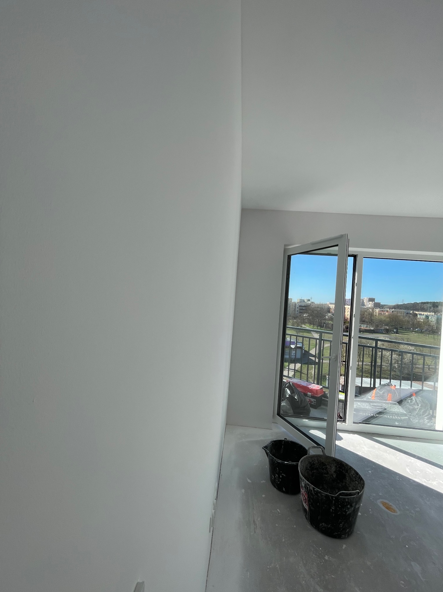 Świeżo pomalowane na biało ściany w pokoju z otwartym oknem balkonowym i dwoma czarnymi wiadrami na podłodze. Widok na miasto w tle.