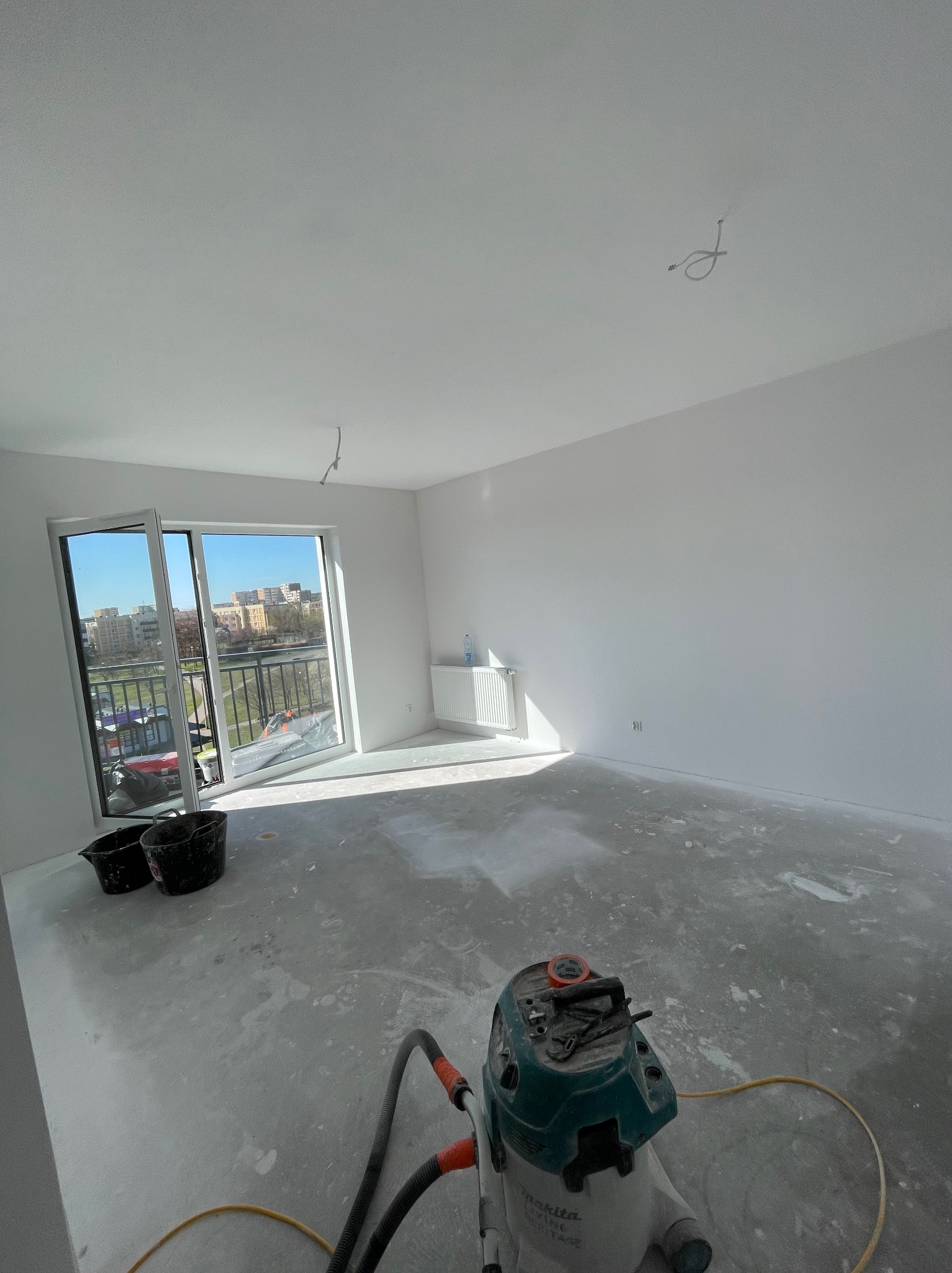 Jasne wnętrze po szpachlowaniu, widoczny odkurzacz przemysłowy i resztki pyłu. Duże okno balkonowe z widokiem na miasto w tle. Świeżo wyszpachlowane ściany i sufit.