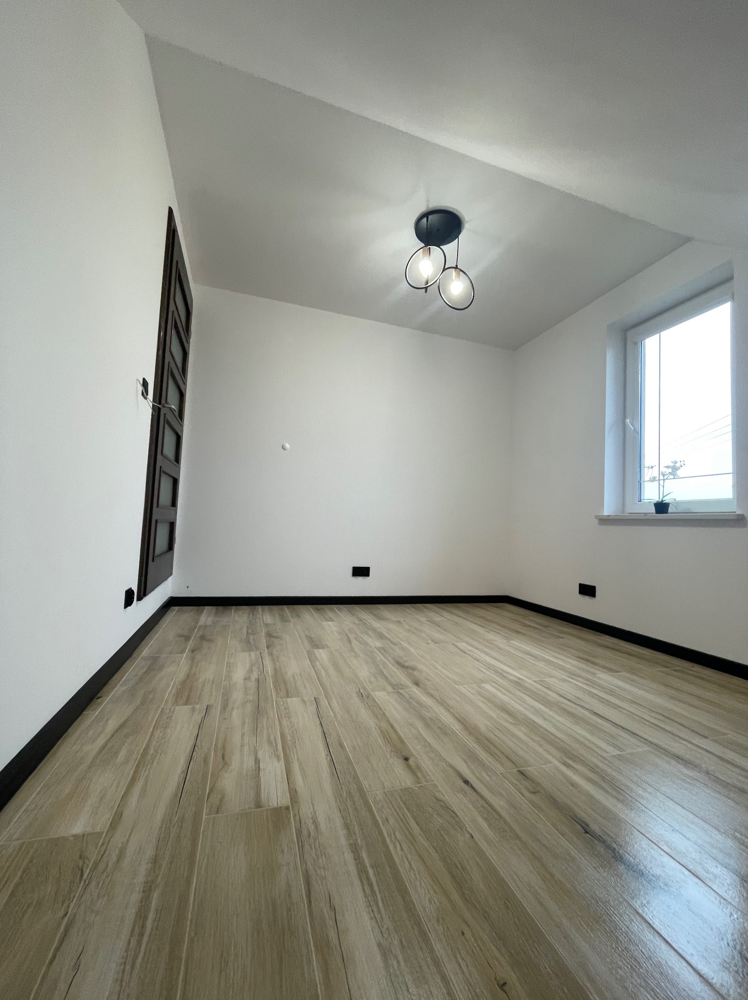 Jasne panele podłogowe w nowym pokoju z białymi ścianami i oknem. Minimalistyczna lampa sufitowa, ciemne listwy przypodłogowe.