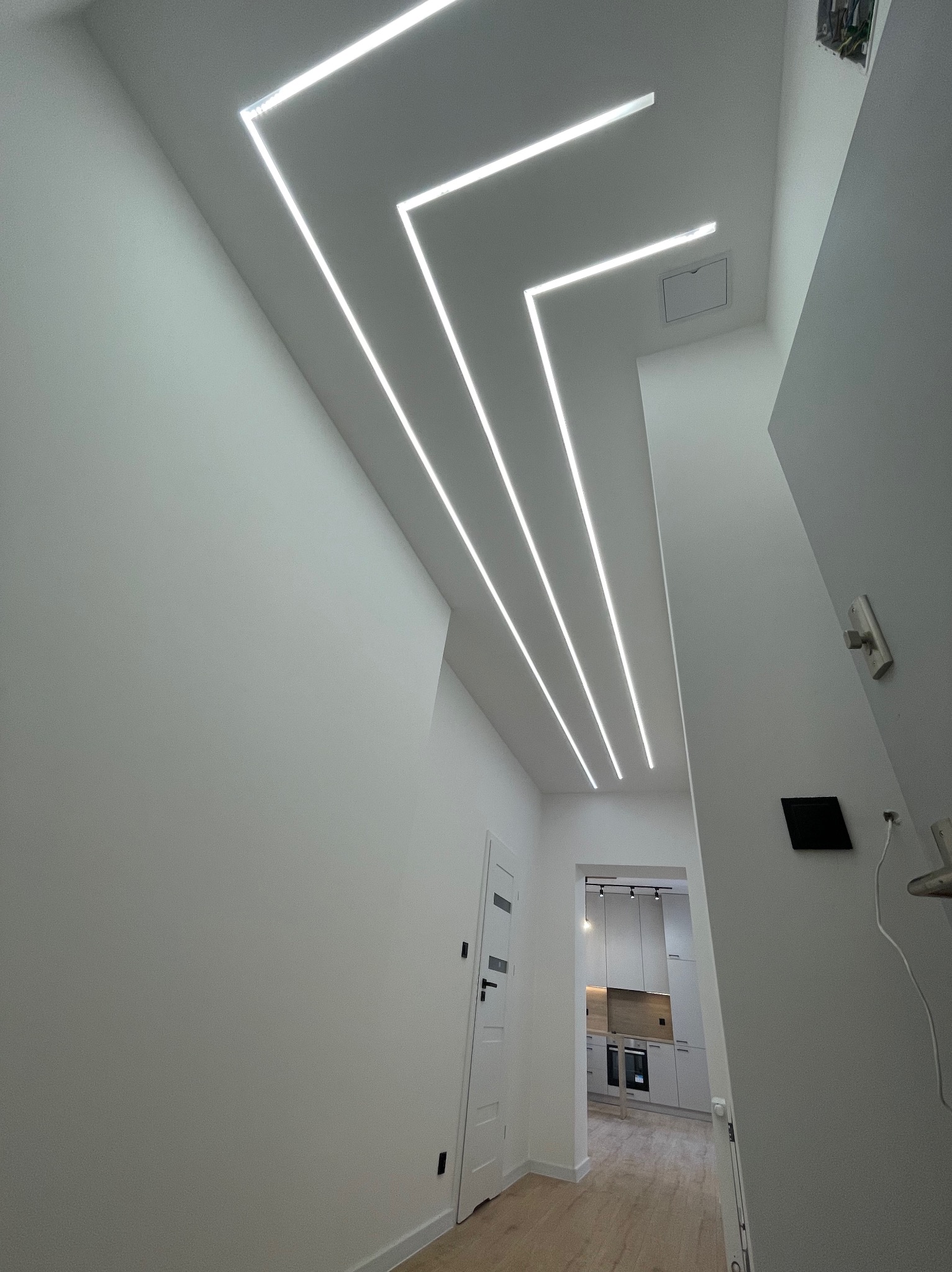 Nowoczesne oświetlenie sufitowe LED w kształcie zygzaka w korytarzu, widok na białą kuchnię w tle. Minimalistyczny design, białe ściany, jasne drewno na podłodze.