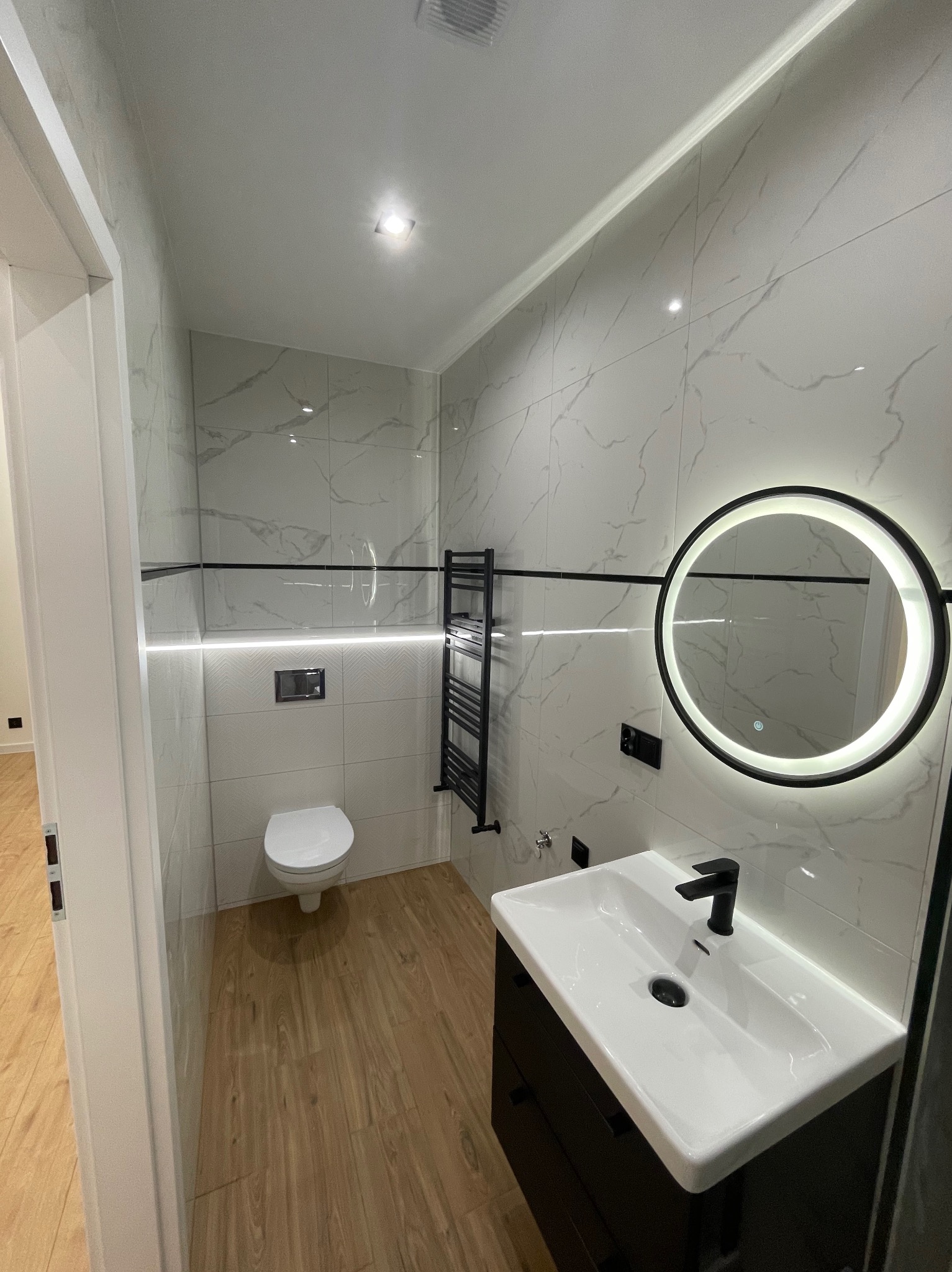 Nowoczesna łazienka po renowacji: białe płytki imitujące marmur, drewnopodobna podłoga, okrągłe lustro z podświetleniem LED i czarne akcenty.