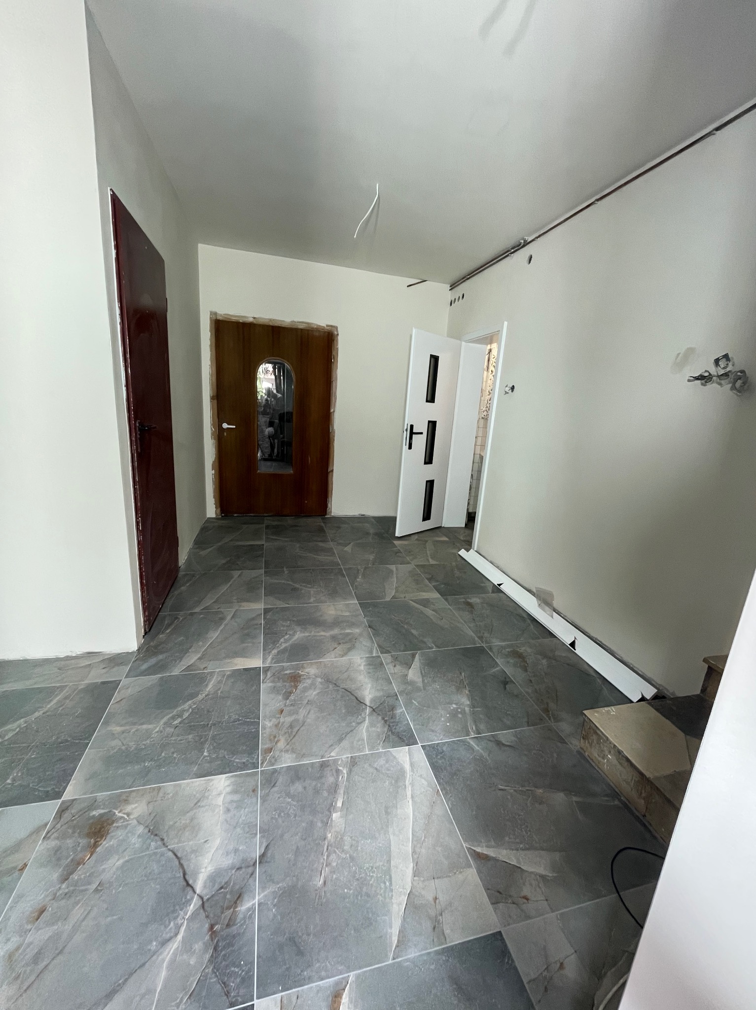 Wykończone wnętrze z szarymi płytkami imitującymi marmur na podłodze, częściowo zamontowane listwy przypodłogowe i dwoje drzwi w różnych kolorach.