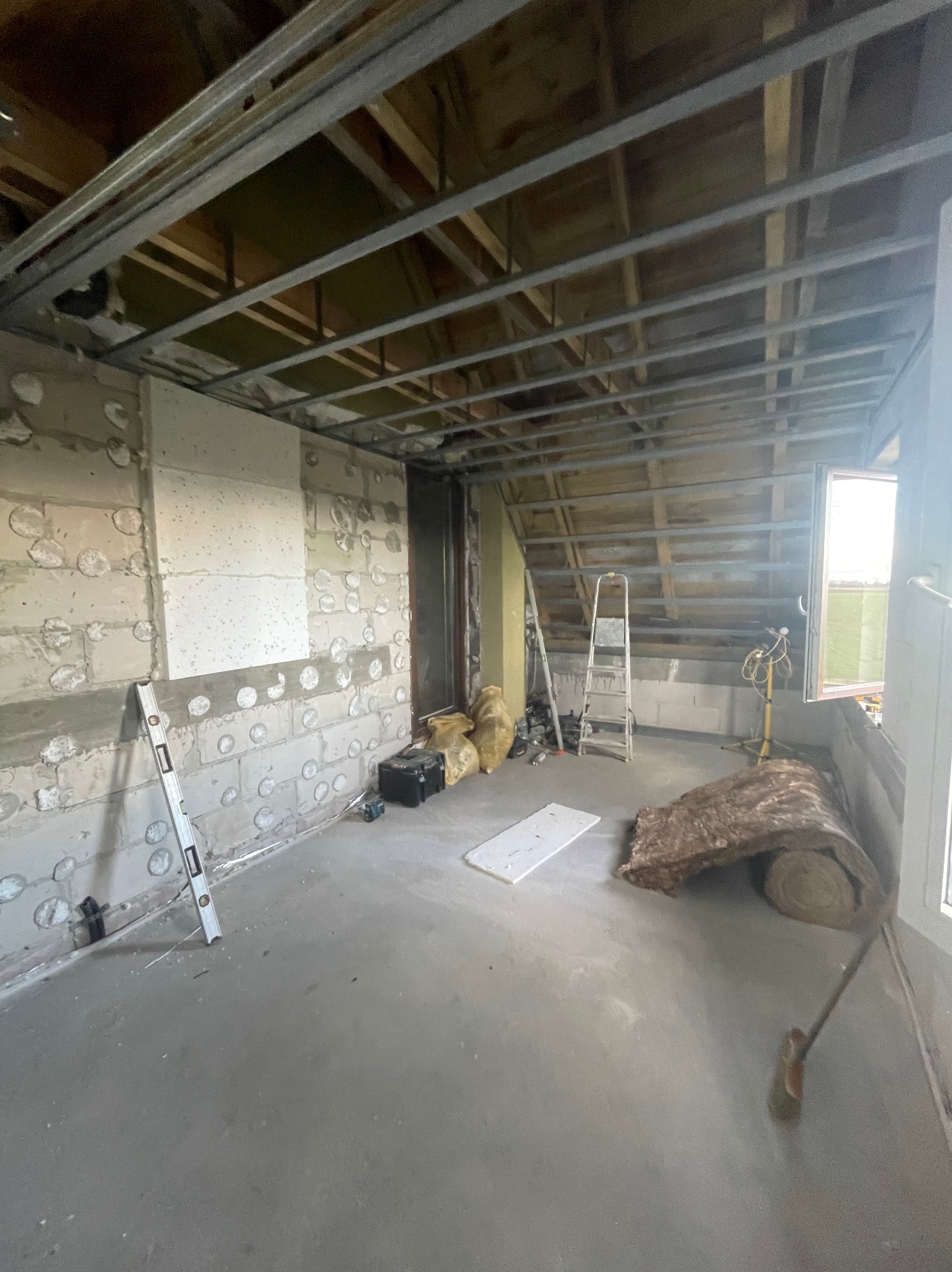 Wnętrze w trakcie prac: surowe ściany z widocznymi bloczkami, częściowo ocieplone, metalowa konstrukcja sufitu podwieszanego, drabina i materiały izolacyjne.