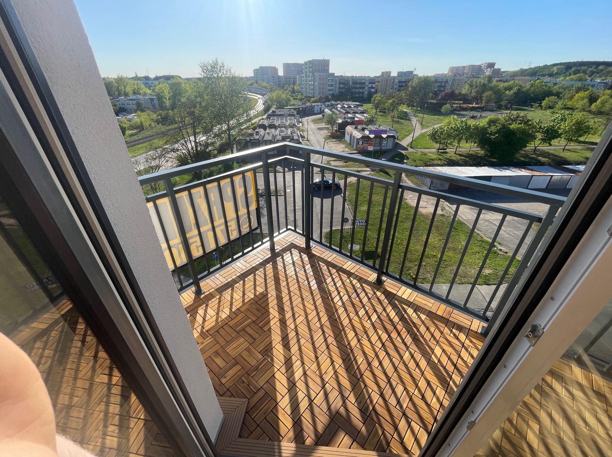 Jasne drewniane płytki na balkonie z metalową balustradą i widokiem na miasto w słoneczny dzień. Charakterystyczne cienie rzucane przez balustradę na podłogę.