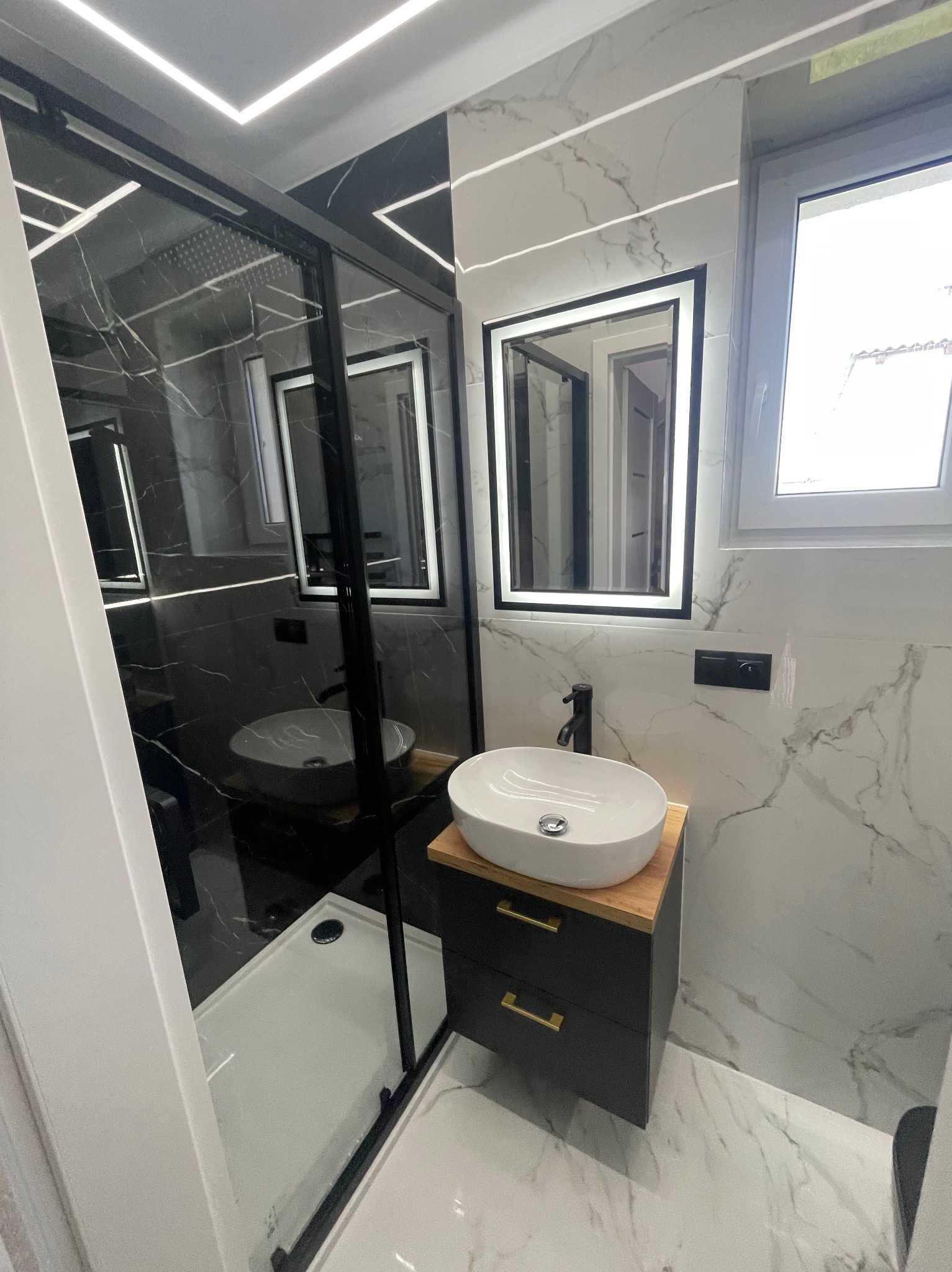 Nowoczesna łazienka po remoncie: kabina prysznicowa typu walk-in z czarnymi profilami, umywalka nablatowa na szafce z drewnianym blatem, marmurowe płytki na ścianach i podłodze.