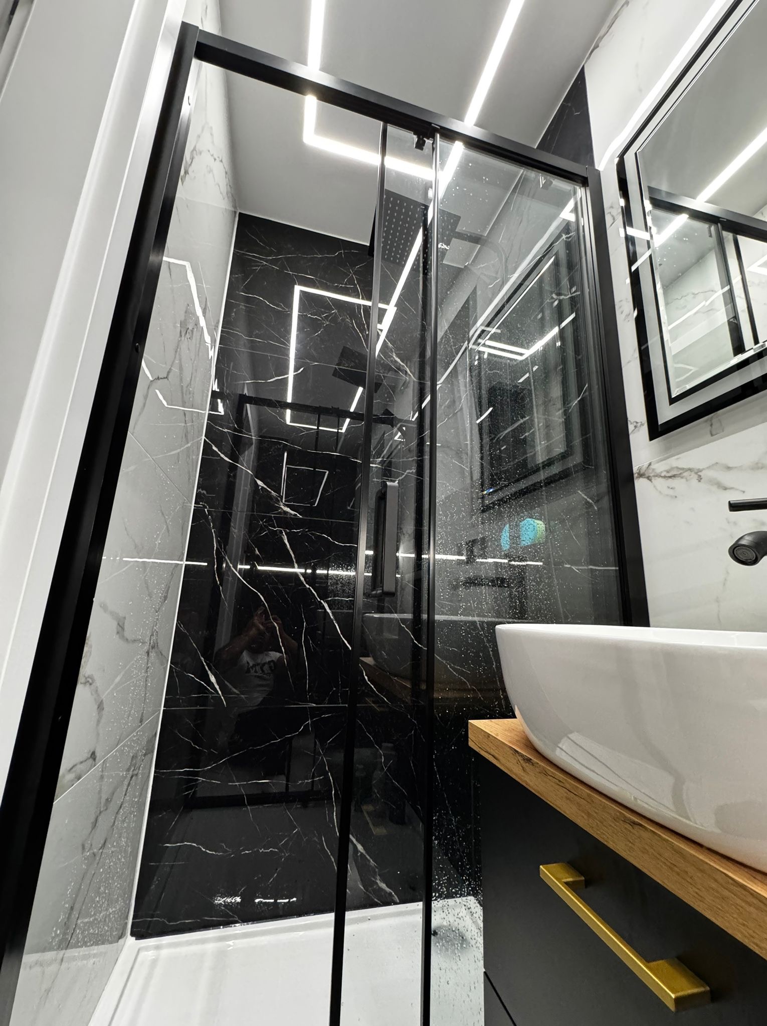 Nowoczesna łazienka z kabiną prysznicową typu walk-in, czarnymi płytkami imitującymi marmur i białym blatem umywalki z drewnianym akcentem. Oświetlenie LED.