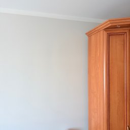 DoMi Group Dominik Mirowski - Jasnoszara ściana z białym gzymsem sufitowym, obok drewnianej szafy narożnej z wbudowanym oświetleniem. Na szafce nocnej lampa solna i budziki.