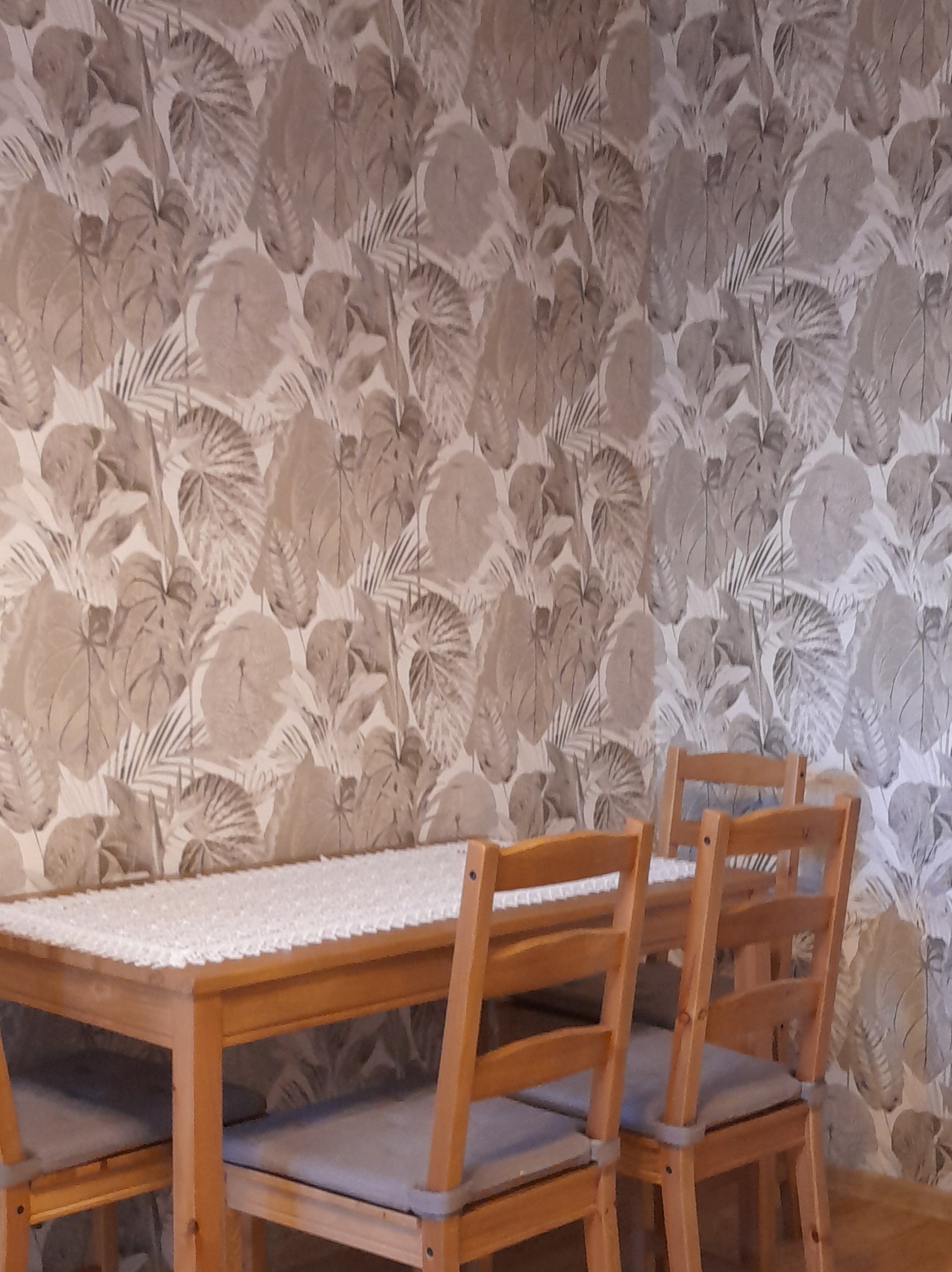 Jadalnia z drewnianym stołem i krzesłami na tle ściany pokrytej tapetą w motywy roślinne.