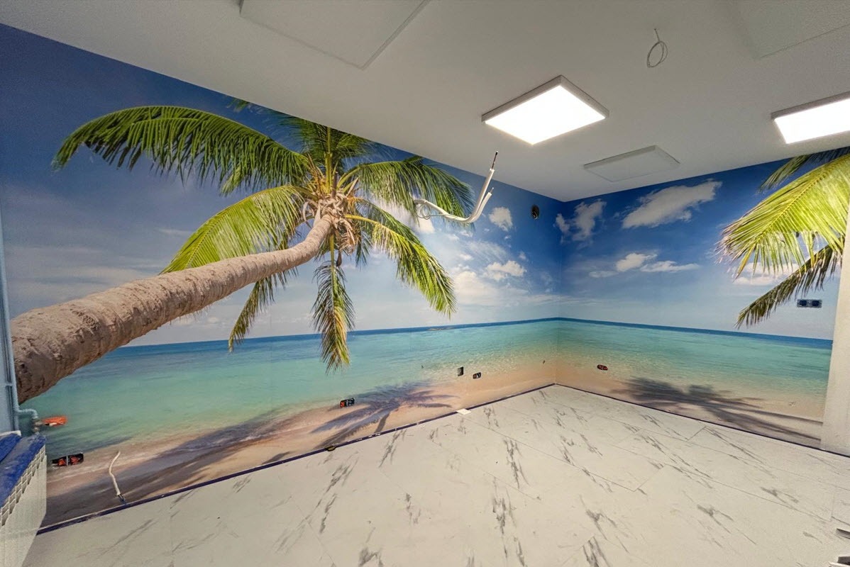 Pokój po tapetowaniu fototapetą z motywem tropikalnej plaży z palmą, błękitnym morzem i niebem. Białe płytki na podłodze i widoczne gniazdka elektryczne.