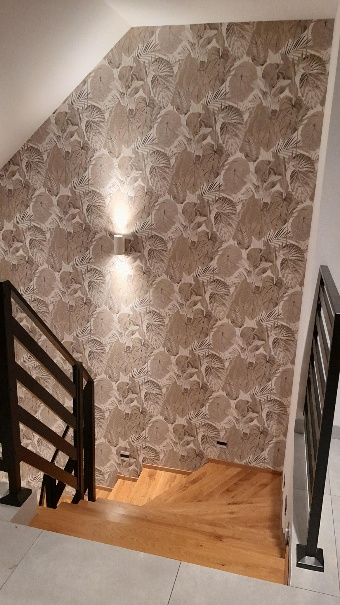 Ściana schodów z tapetą w liście, oświetlona kinkietami. Drewniane schody i czarna balustrada. Widok z góry, neutralna kolorystyka.