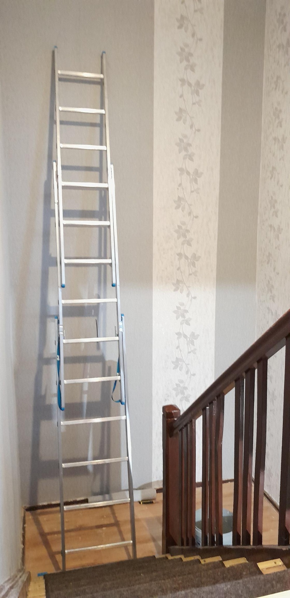 Aluminiowa drabina oparta o ścianę z paskami: gładka szara i wzorzysta w delikatne kwiaty. Widoczne schody i drewniana balustrada. Tapeta w trakcie montażu.