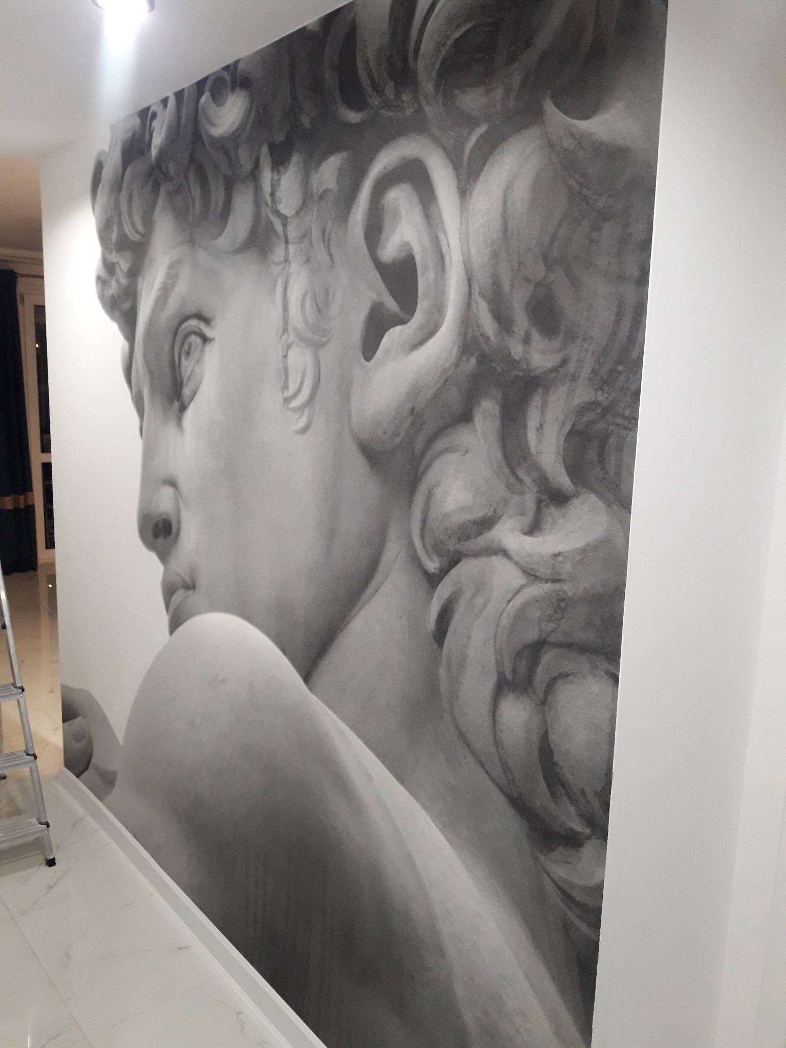 Ściana z fototapetą przedstawiającą popiersie Dawida w odcieniach szarości. Widoczny fragment drabiny i marmurowa podłoga. Ujęcie z dołu, podkreślające monumentalność.