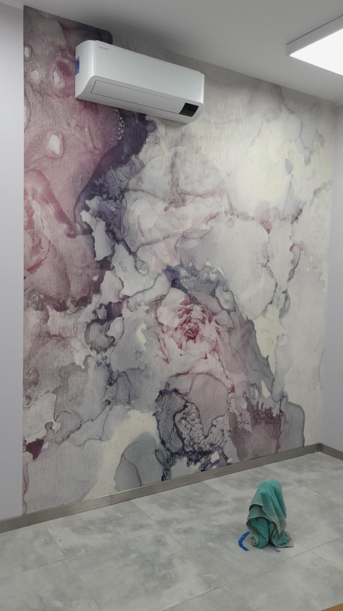 Ściana po tapetowaniu z wzorem akwareli w odcieniach różu i szarości, pod klimatyzatorem. Na podłodze leży zwinięty ręcznik.