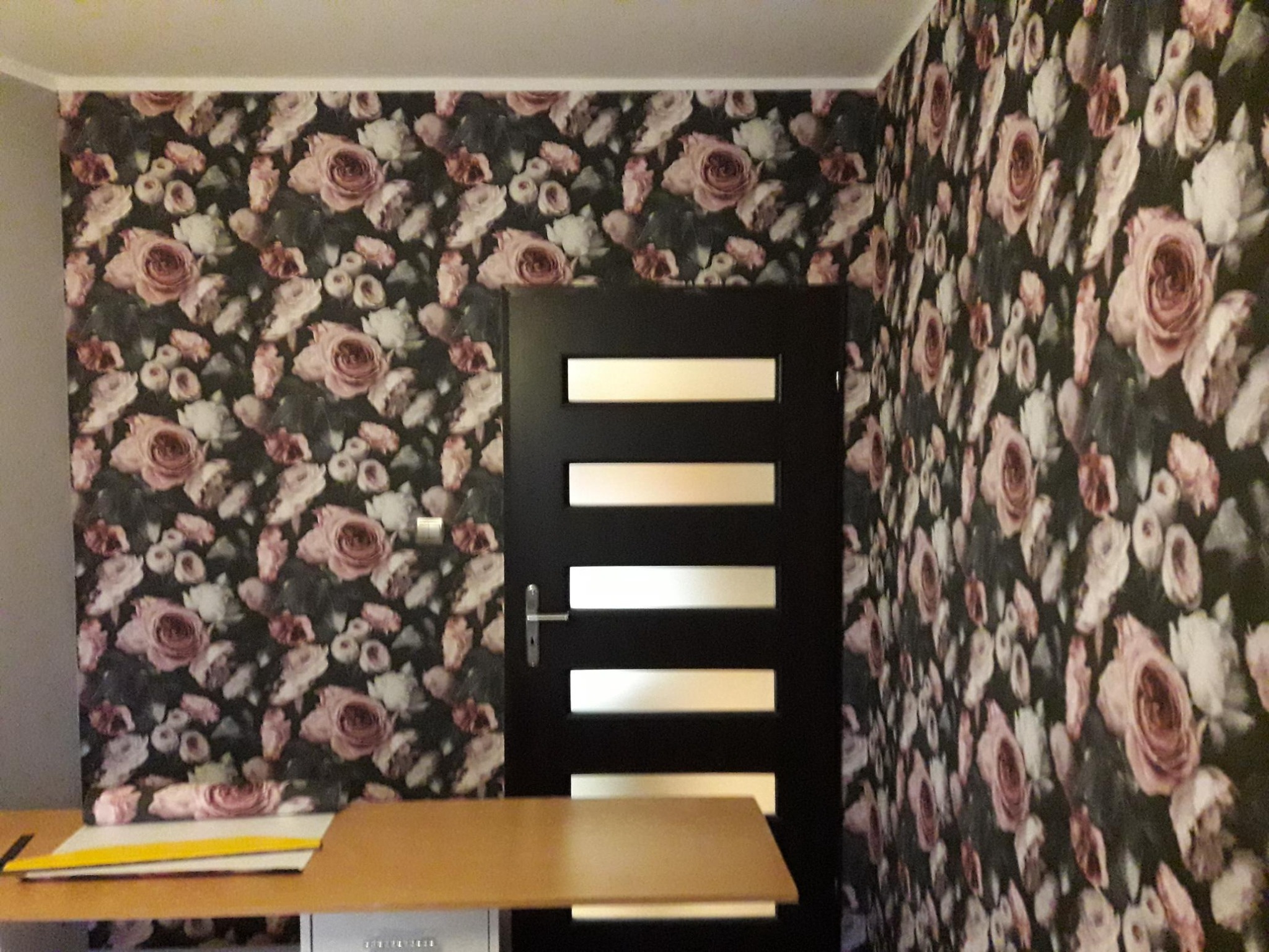 Ściana w ciemne, kwiatowe tapety z motywem róż, sąsiadująca z czarnymi drzwiami z poziomymi, jasnymi wstawkami, widziana z perspektywy niskiego kąta.