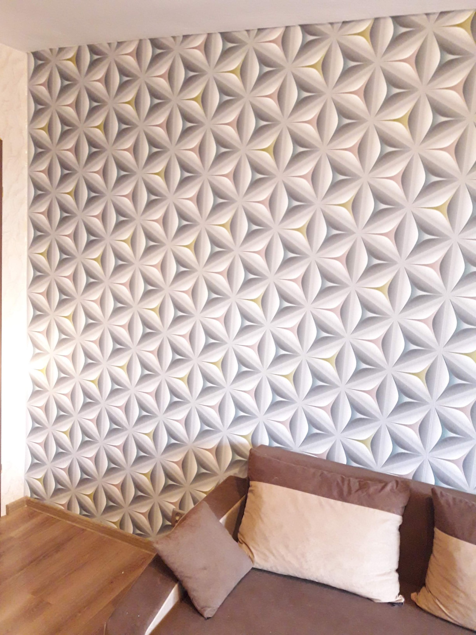 Ściana z tapetą o geometrycznym wzorze kwiatów w odcieniach szarości, bieli i żółci, widoczna nad brązową sofą z poduszkami w pokoju.