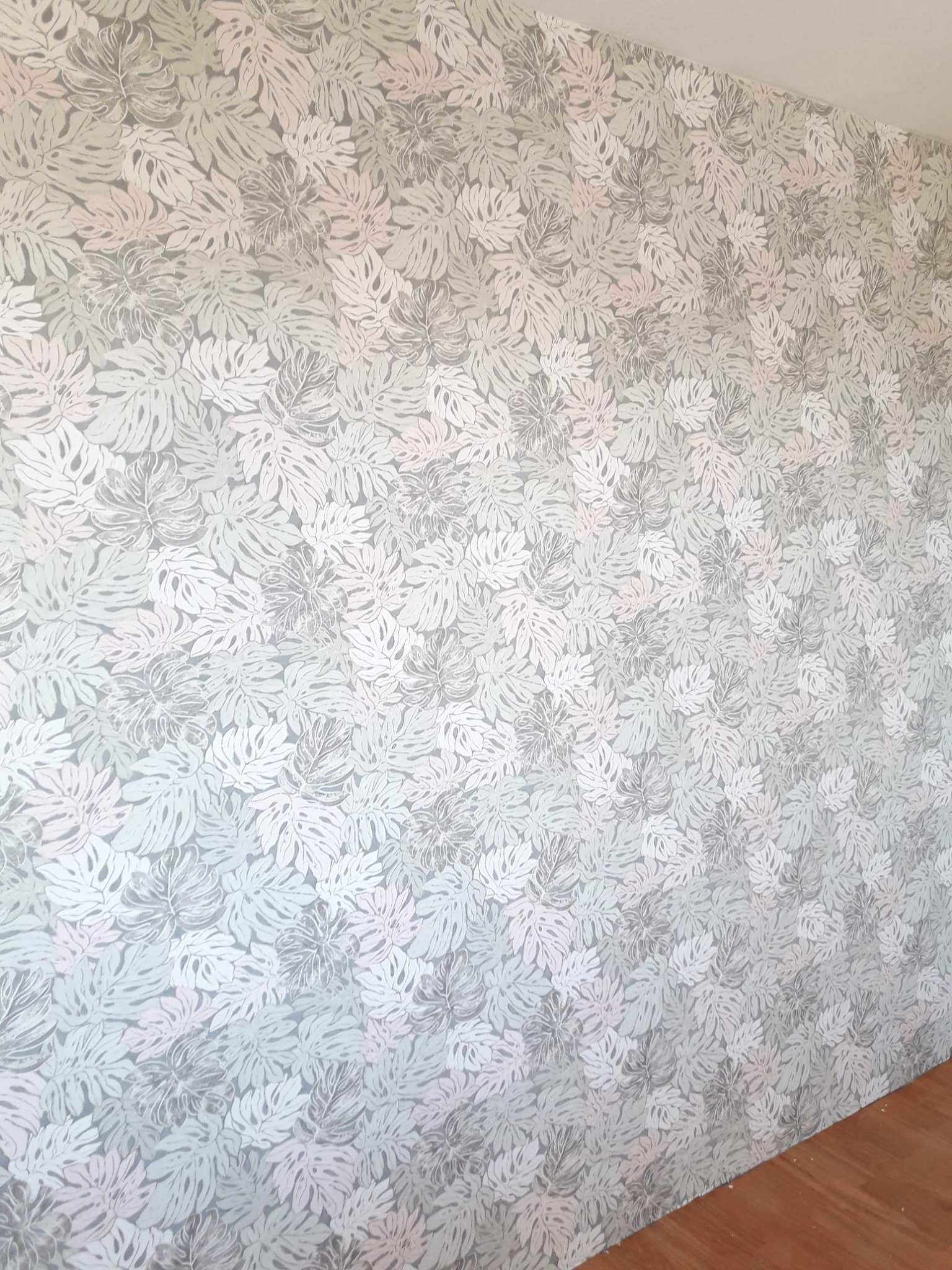 Ściana pokryta tapetą w delikatny wzór liści monstery w odcieniach szarości i różu, widoczny fragment drewnianej podłogi.