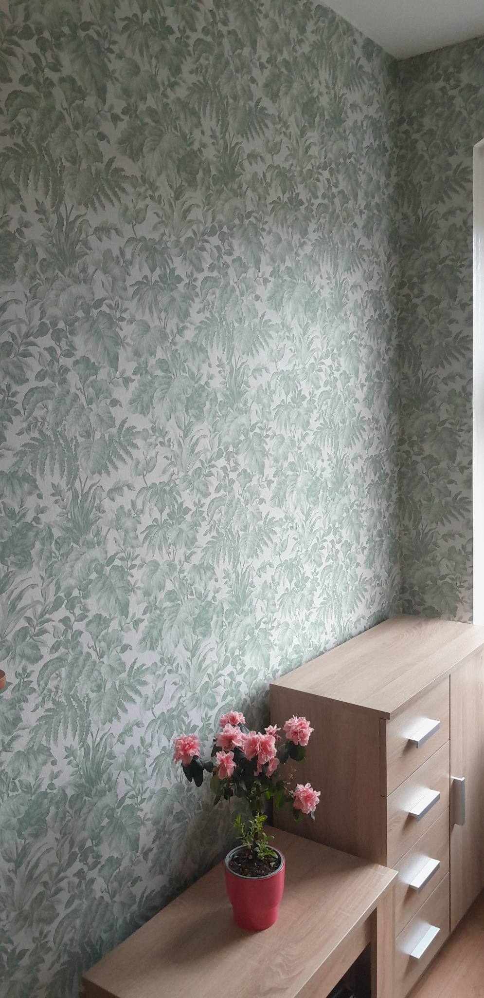 Ściana pokryta tapetą w roślinny wzór w odcieniach zieleni i bieli. Na komodzie stoi doniczka z różowymi kwiatami. Jasne wnętrze.