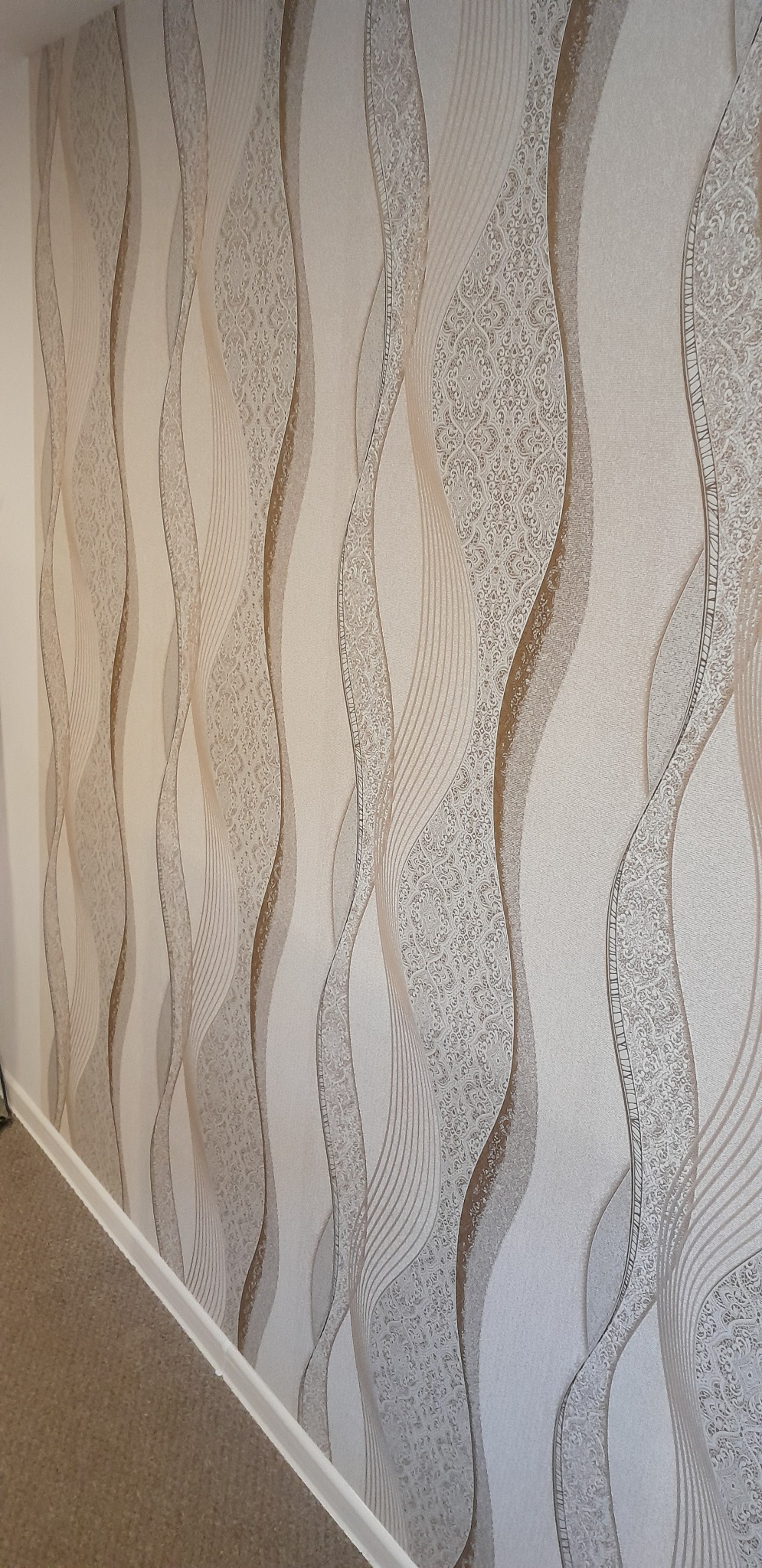 Ściana z beżową tapetą w faliste wzory, imitujące tkaninę. Widoczny fragment podłogi i listwy przypodłogowej. Delikatne światło podkreśla fakturę tapety.