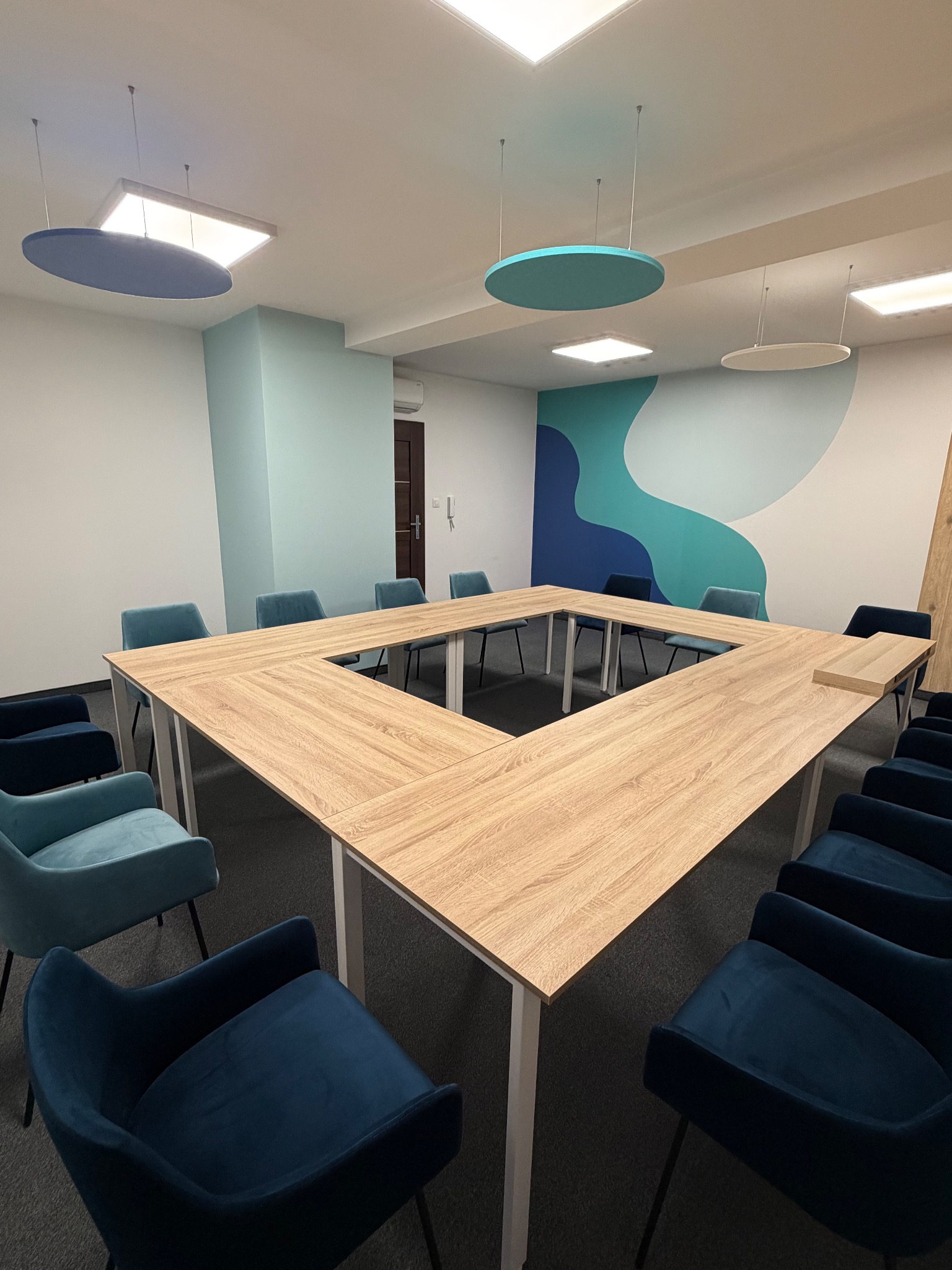 Nowoczesna sala konferencyjna z drewnianym stołem w kształcie kwadratu i niebieskimi krzesłami. Na ścianie dekoracyjny wzór w odcieniach błękitu i granatu. Oświetlenie sufitowe.