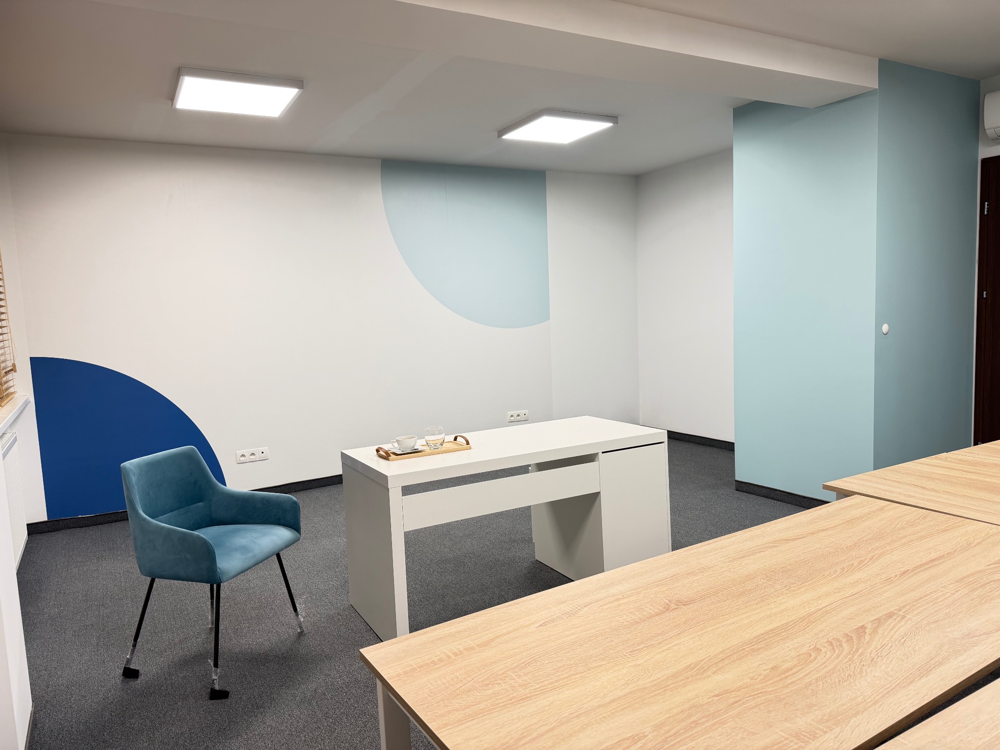 Błękitne akcenty w nowoczesnym biurze: ściana z półokręgiem, krzesło i biurko. Jasne wnętrze z drewnianymi blatami i szarą wykładziną. Minimalistyczny design.