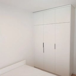 Dariusz Zsuiard - Biała, minimalistyczna szafa wnękowa z czterema drzwiami i długimi, smukłymi uchwytami, dopasowana do białych ścian i listwy przypodłogowej, z fragmentem łóżka w dolnej części kadru.