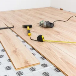 Proces układania paneli podłogowych: widoczne narzędzia, podkład z nadrukiem i fragment ułożonej podłogi z jasnego drewna.