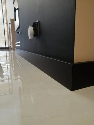 Czarne listwy przypodłogowe zamontowane na styku ciemnej ściany i jasnej, błyszczącej podłogi, widoczny biały czujnik na ścianie.