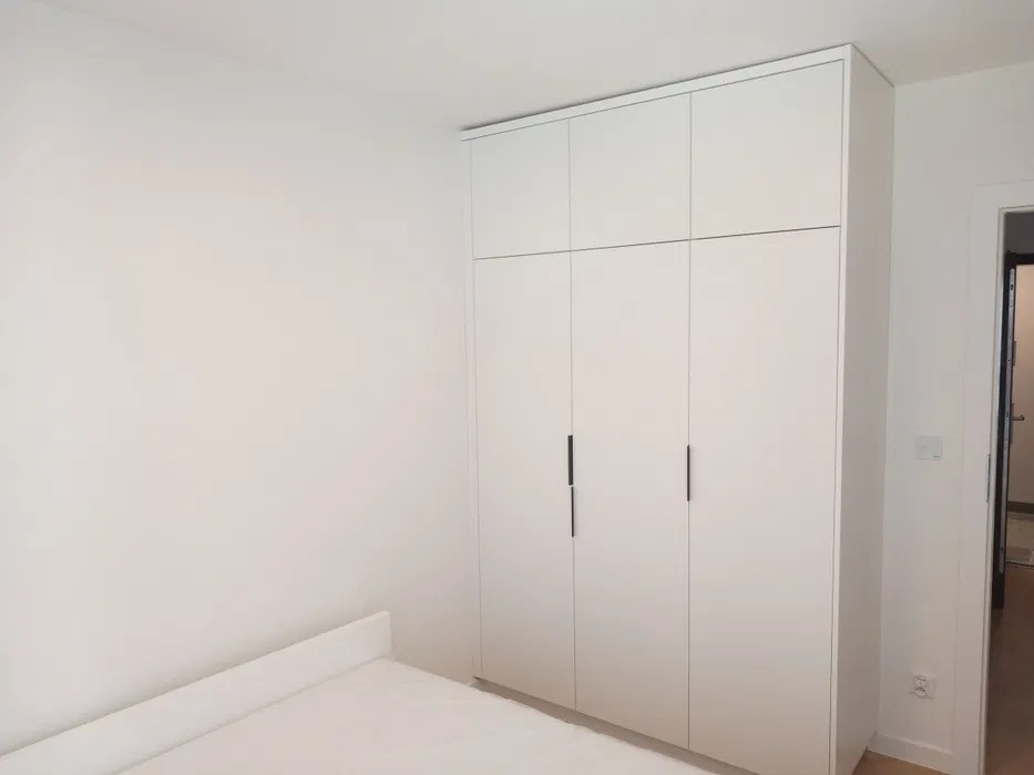 Biała, minimalistyczna szafa wnękowa z czterema drzwiami i długimi, smukłymi uchwytami, dopasowana do białych ścian i listwy przypodłogowej, z fragmentem łóżka w dolnej części kadru.